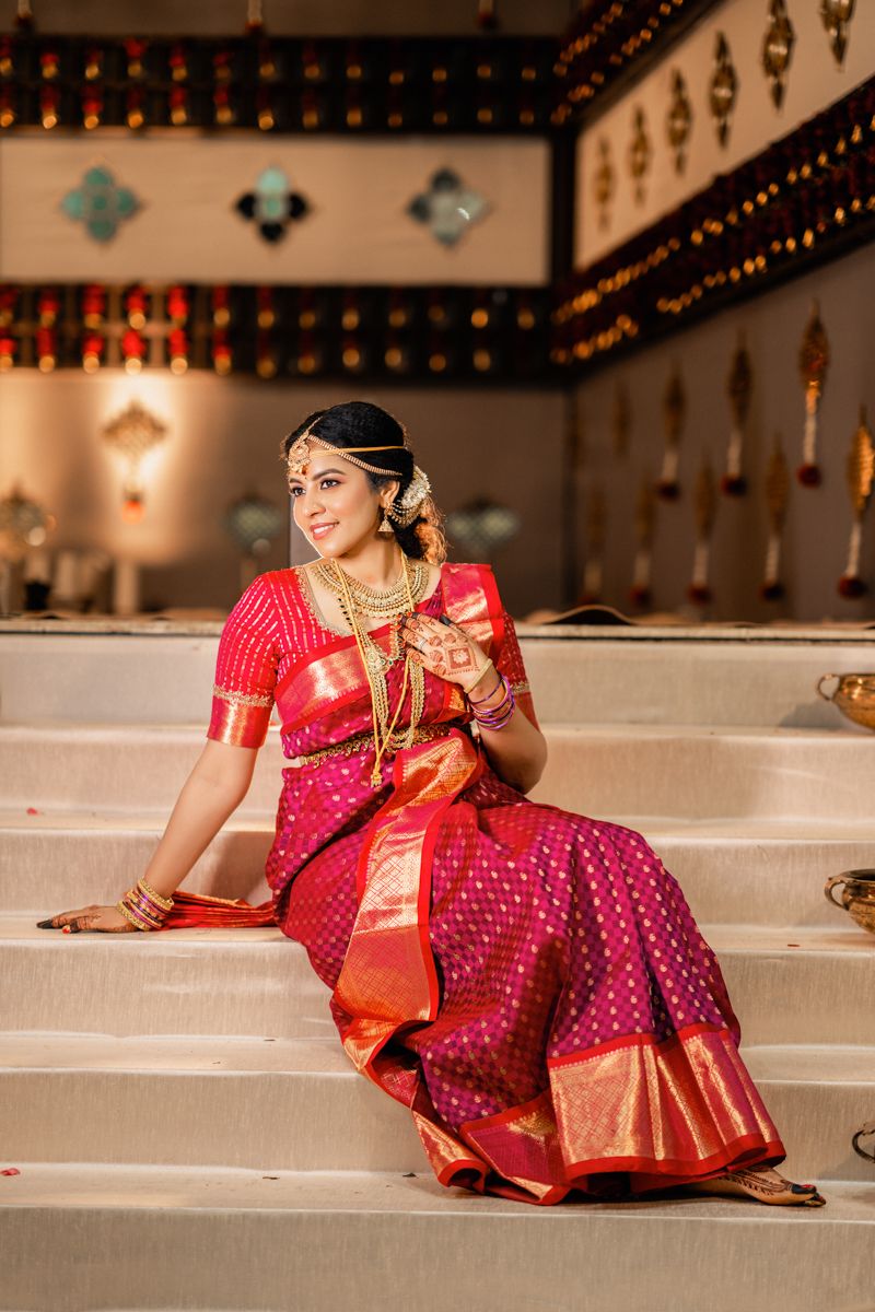 South Asian bride in vibrant red silk saree with gold jewelry sits gracefully on temple steps.