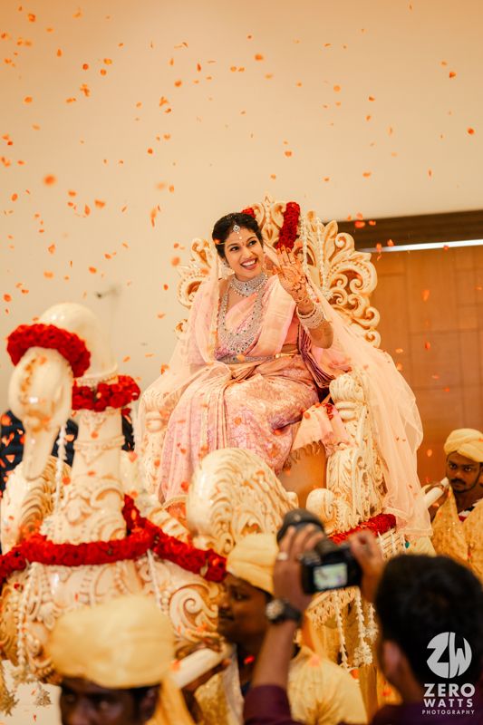 A joyful bride in a pink and silver sari sits on an ornate throne during a traditional Indian wedding ceremony.