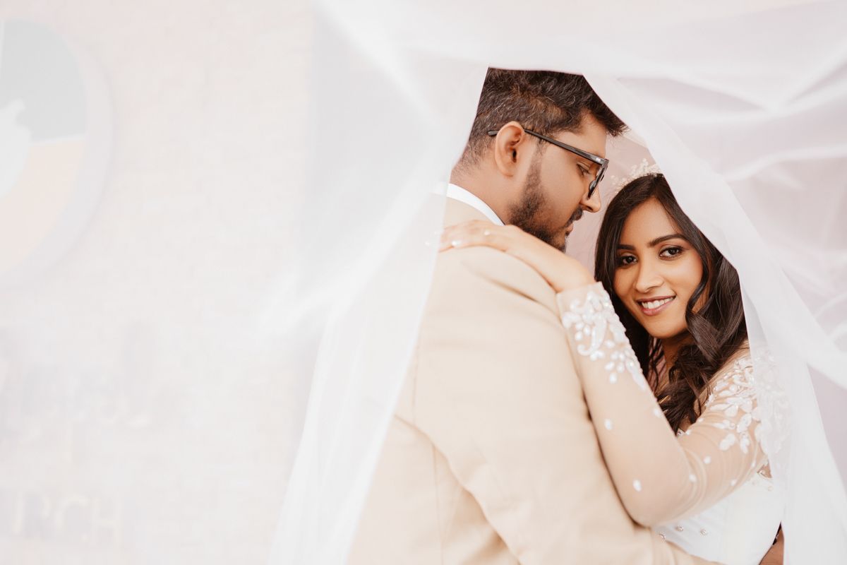 A couple shares an intimate moment under sheer white fabric against a soft light background.