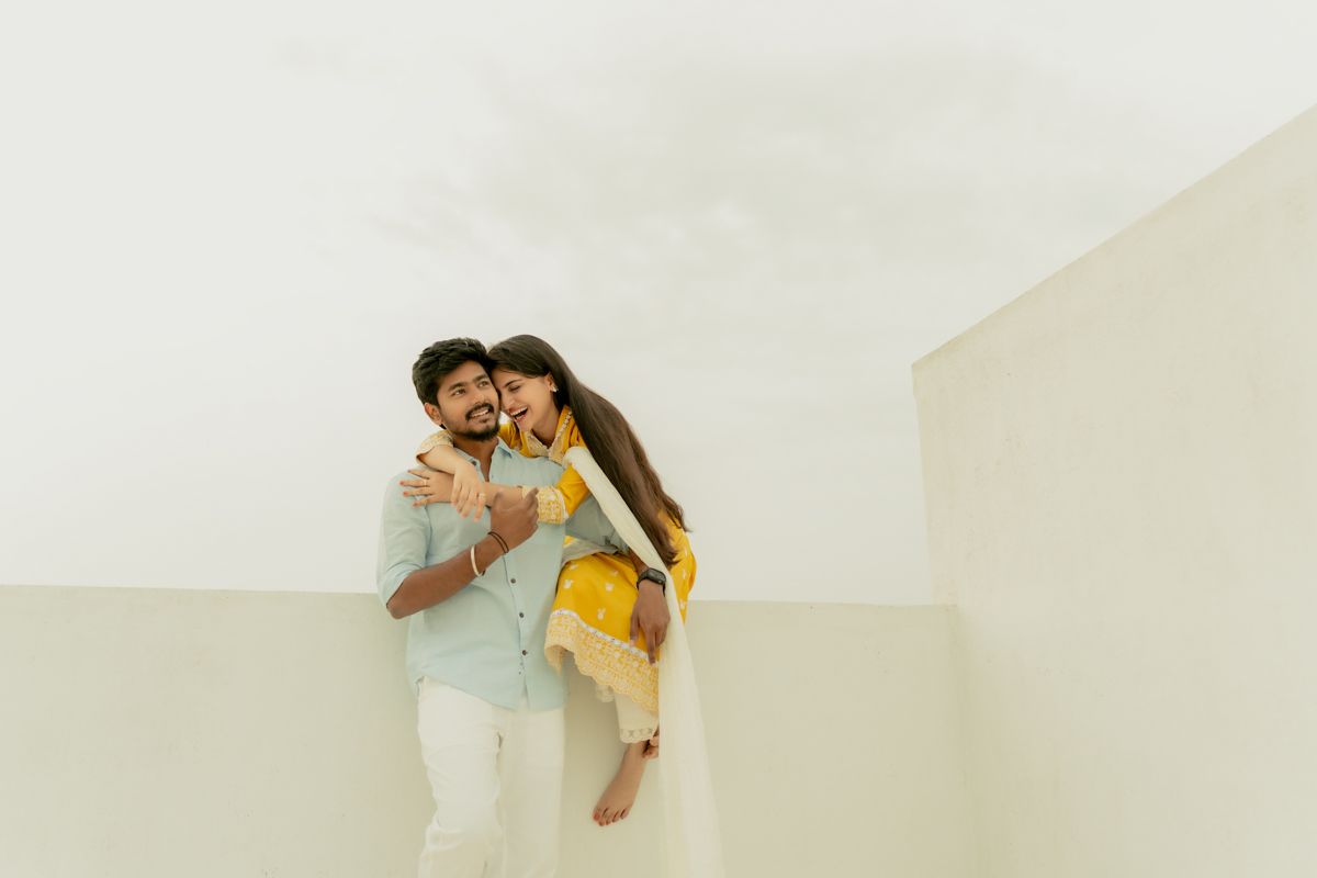 A couple embraces against a minimalist white wall while dressed in yellow and light blue outfits.