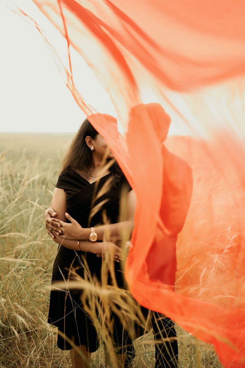 Person in a black dress stands in a wheat field with flowing coral fabric billowing dramatically in the wind.