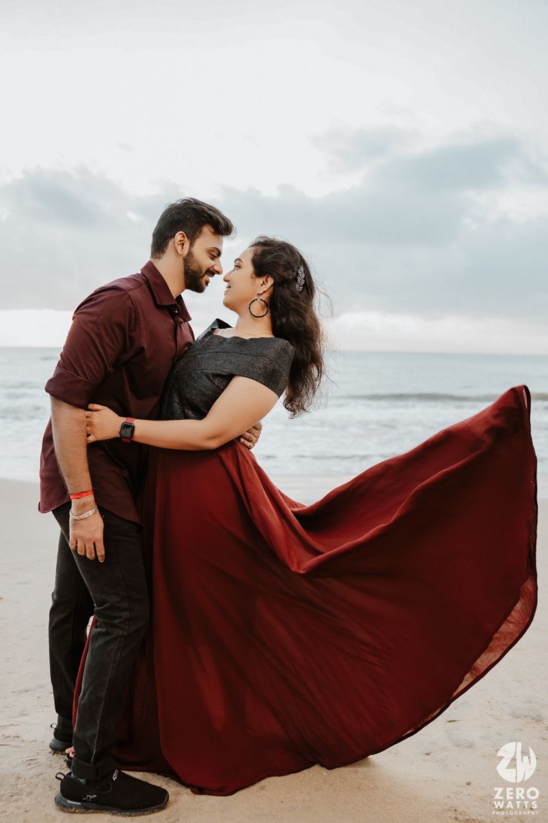 A couple shares an intimate dance pose on the beach with flowing red dress catching the wind.