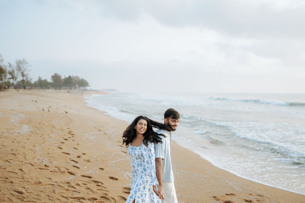 Couple walking along sandy beach shore with waves and cloudy sky.