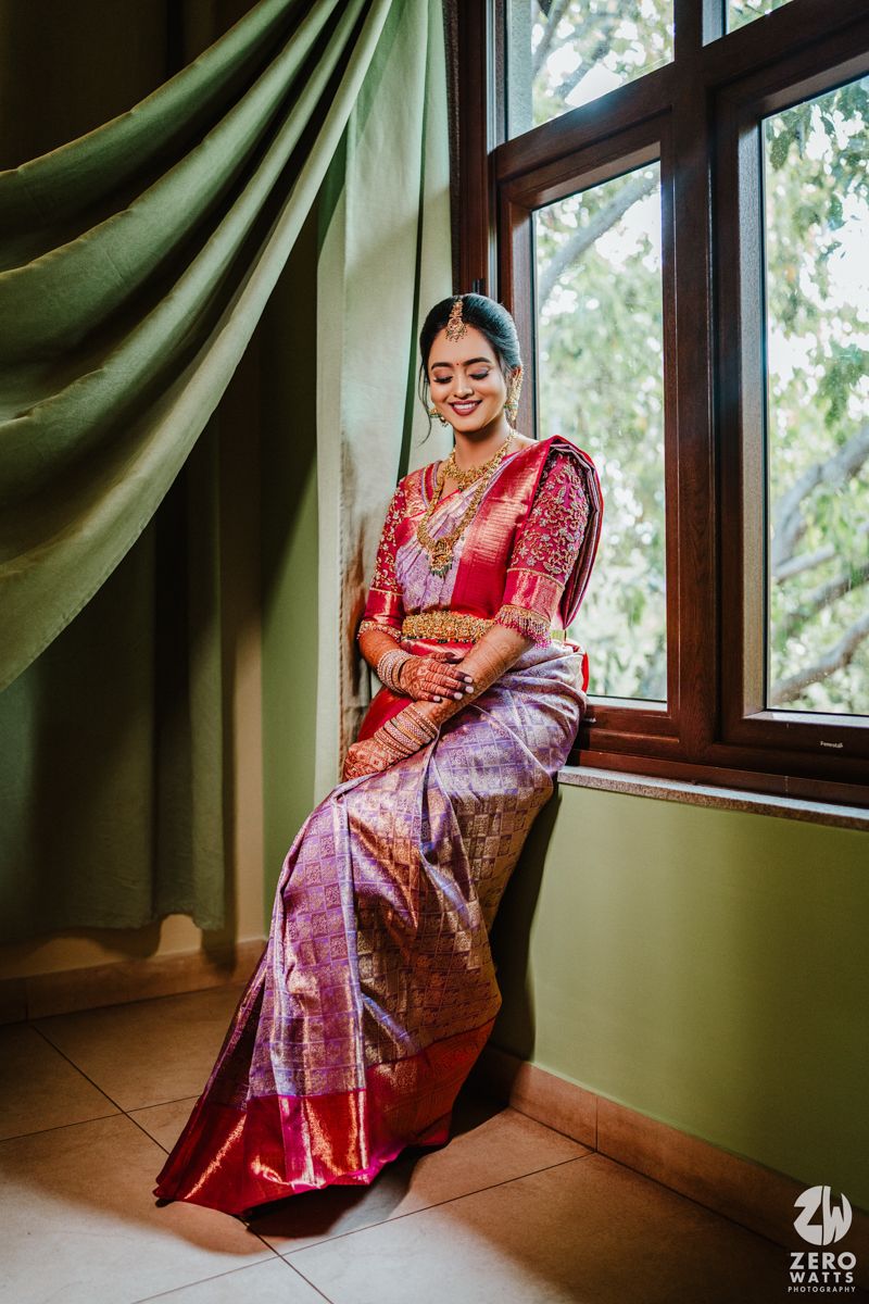 A person in a red and purple silk saree poses elegantly near a window with green drapes in natural lighting.