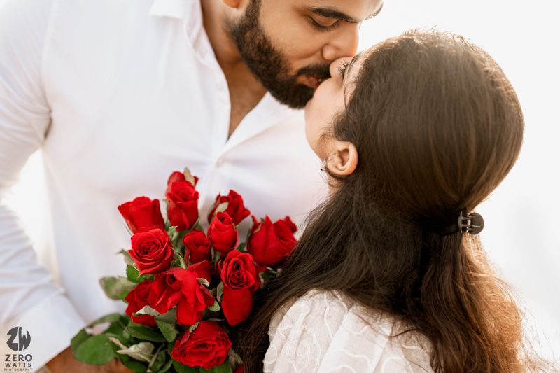 A romantic couple embraces on a rocky beach with red roses and white outfits against an ocean backdrop.