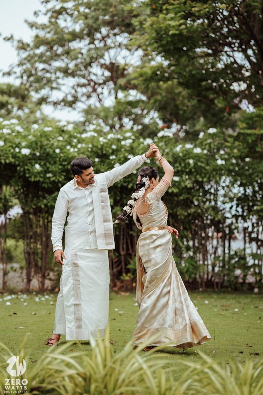 Romantic couple in traditional white outfits dancing together playfully in a lush green outdoor garden setting.