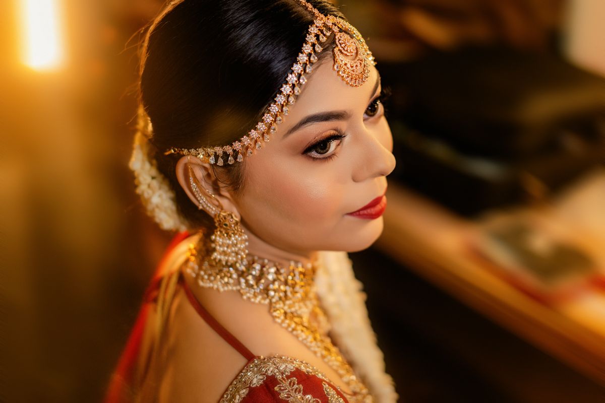 Side profile of bride wearing traditional Indian wedding jewelry and makeup in warm ambient lighting.