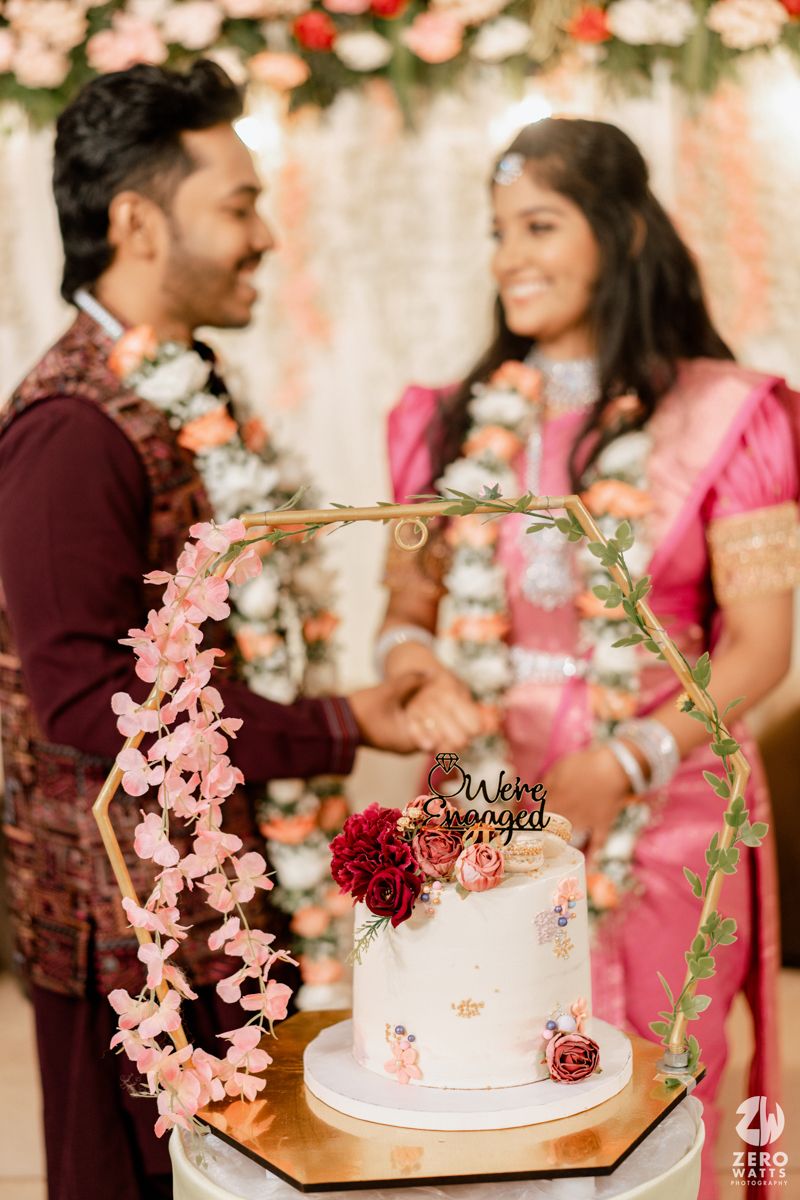 An elegant wedding cake with pink flowers and golden arch stands in foreground while a couple celebrates behind it.