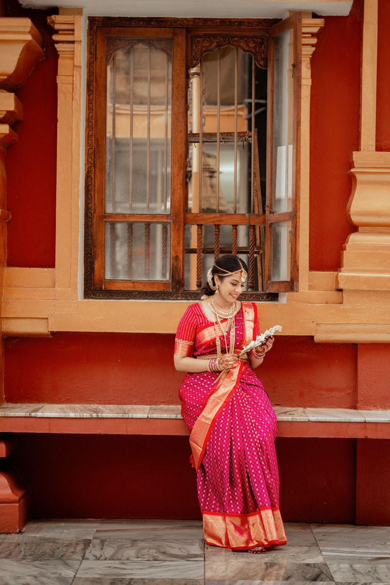 A person in a vibrant red and gold silk saree checks their phone while standing beside a vintage window.