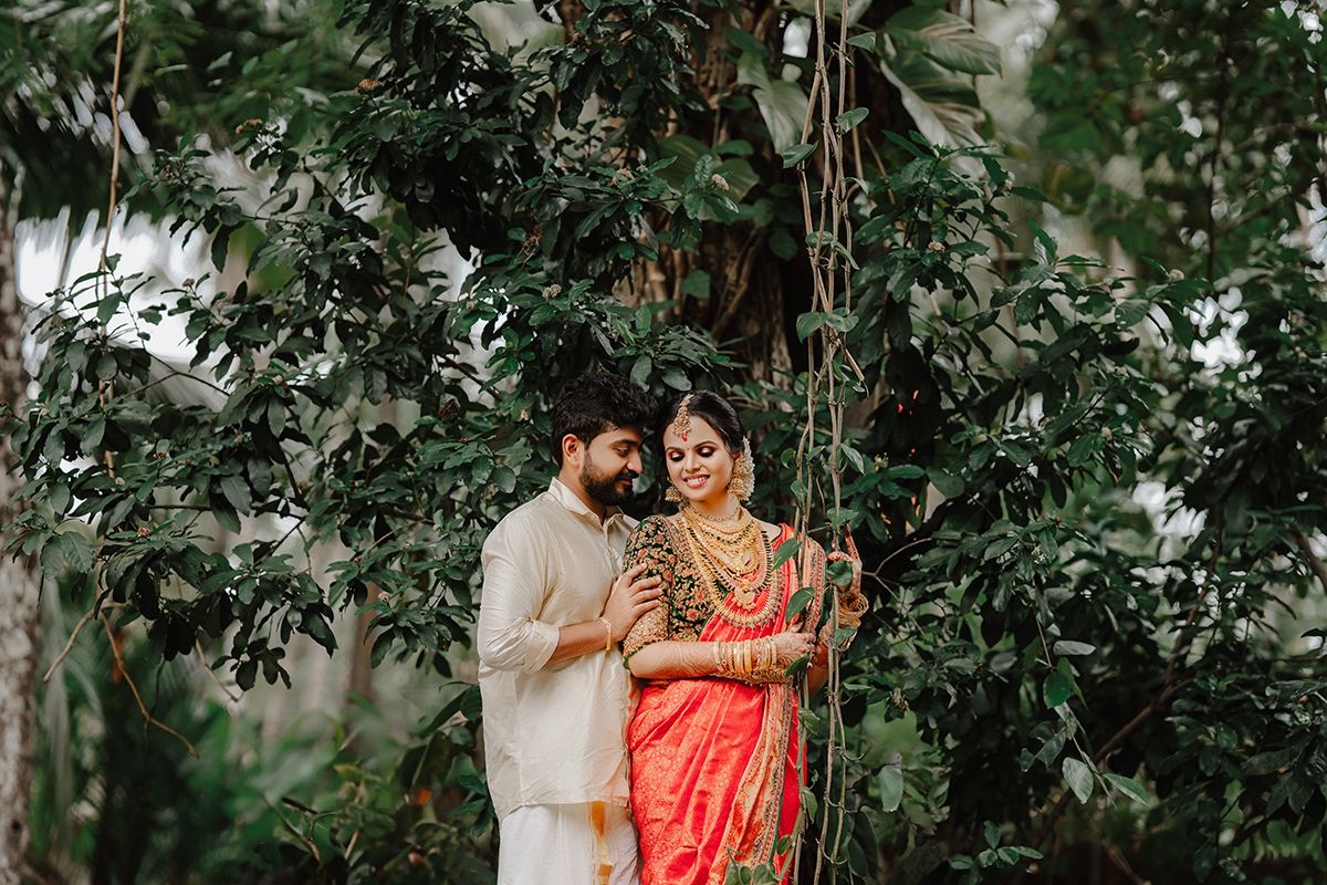 A couple in traditional Indian wedding attire embrace under lush green foliage during an outdoor photoshoot.