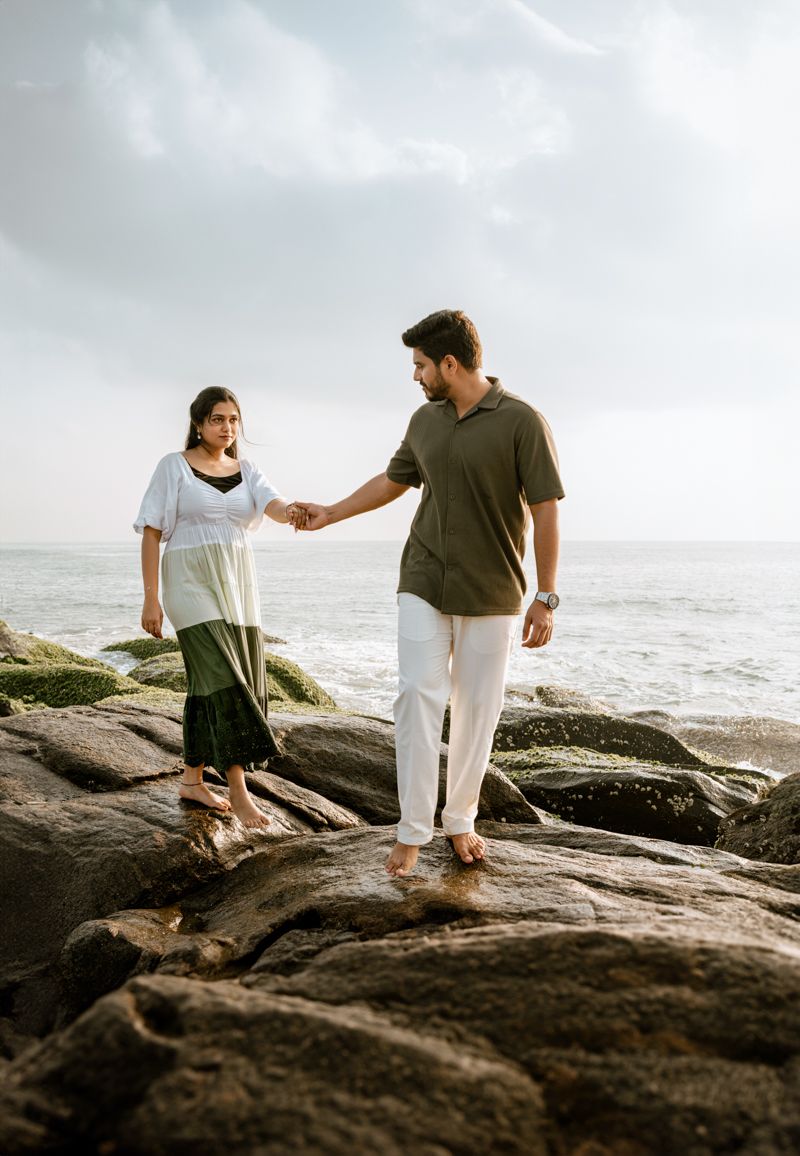 Couple holding hands while balancing on coastal rocks near ocean waves.