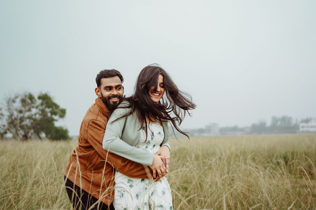 Playful couple embracing and laughing together in a grassy field on cloudy day.