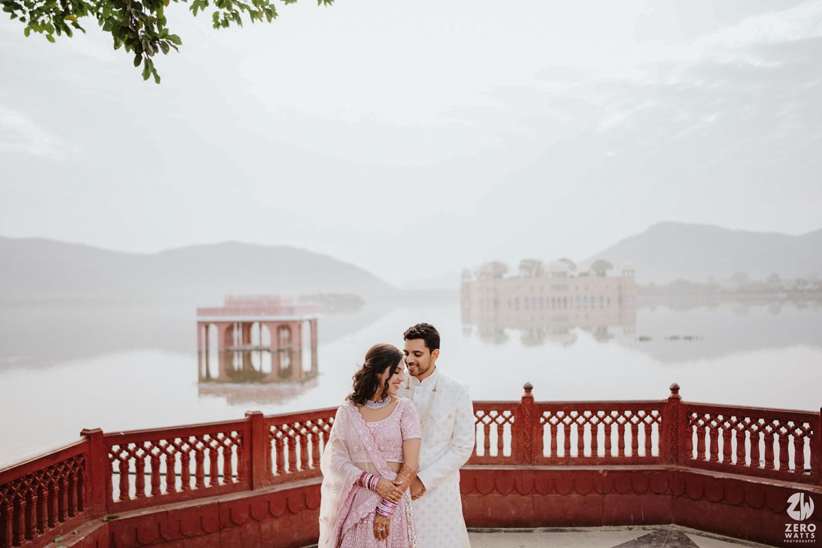 A couple stands on a red-walled terrace overlooking a misty lake with ornate structures in the background.