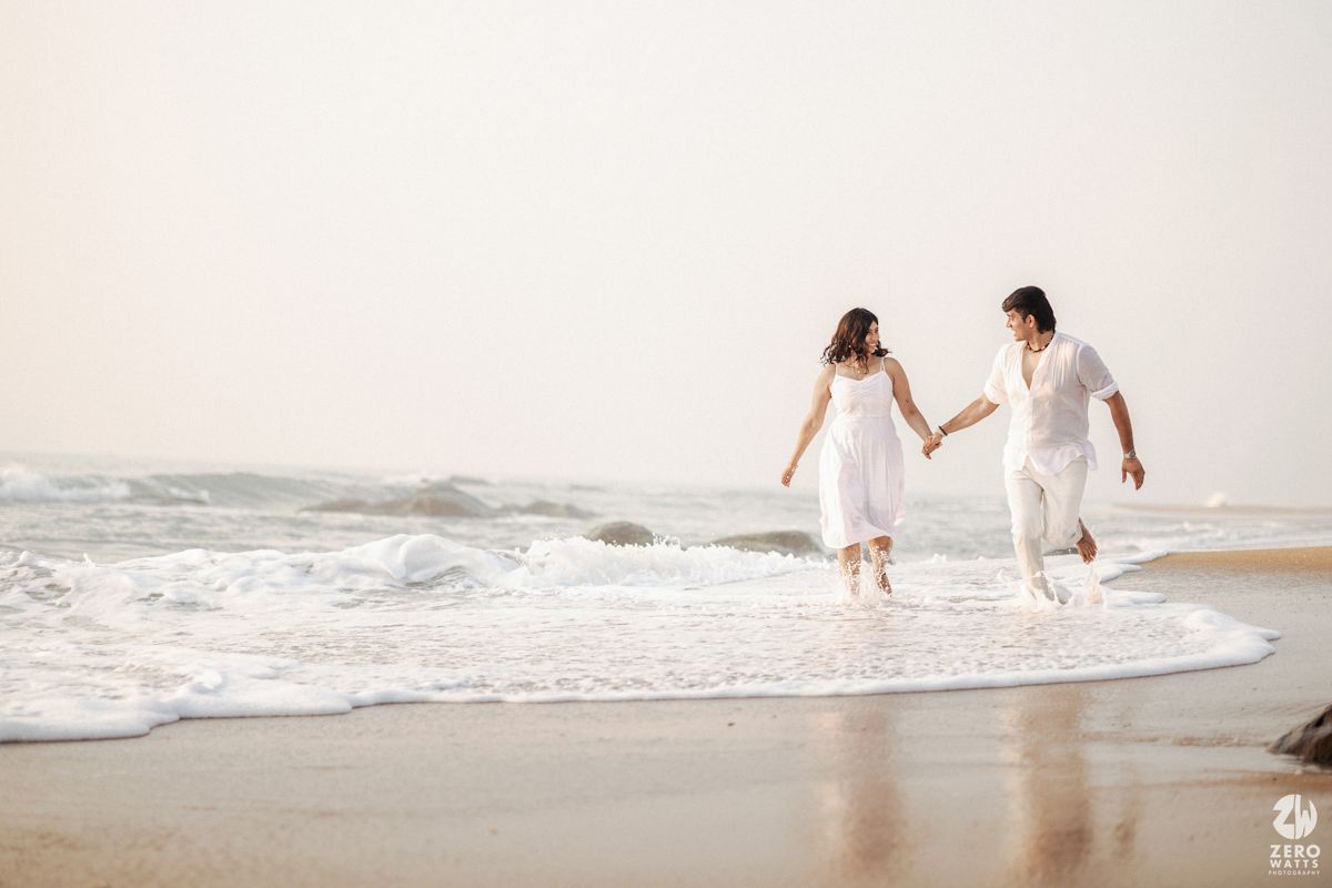 A couple in white outfits holding hands while running along a misty beach at sunset.