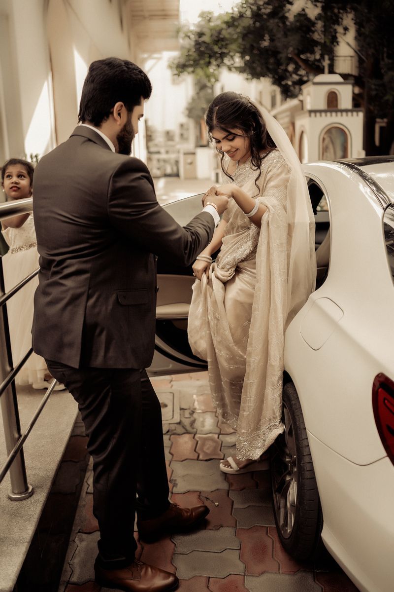 A romantic wedding moment captured as a groom helps his bride step out of a white car while wearing traditional attire.