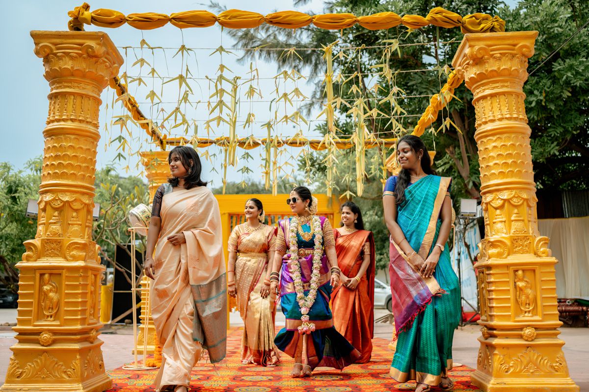A group in vibrant sarees gather under a decorated golden mandap arch with hanging floral decorations at an outdoor ceremony.