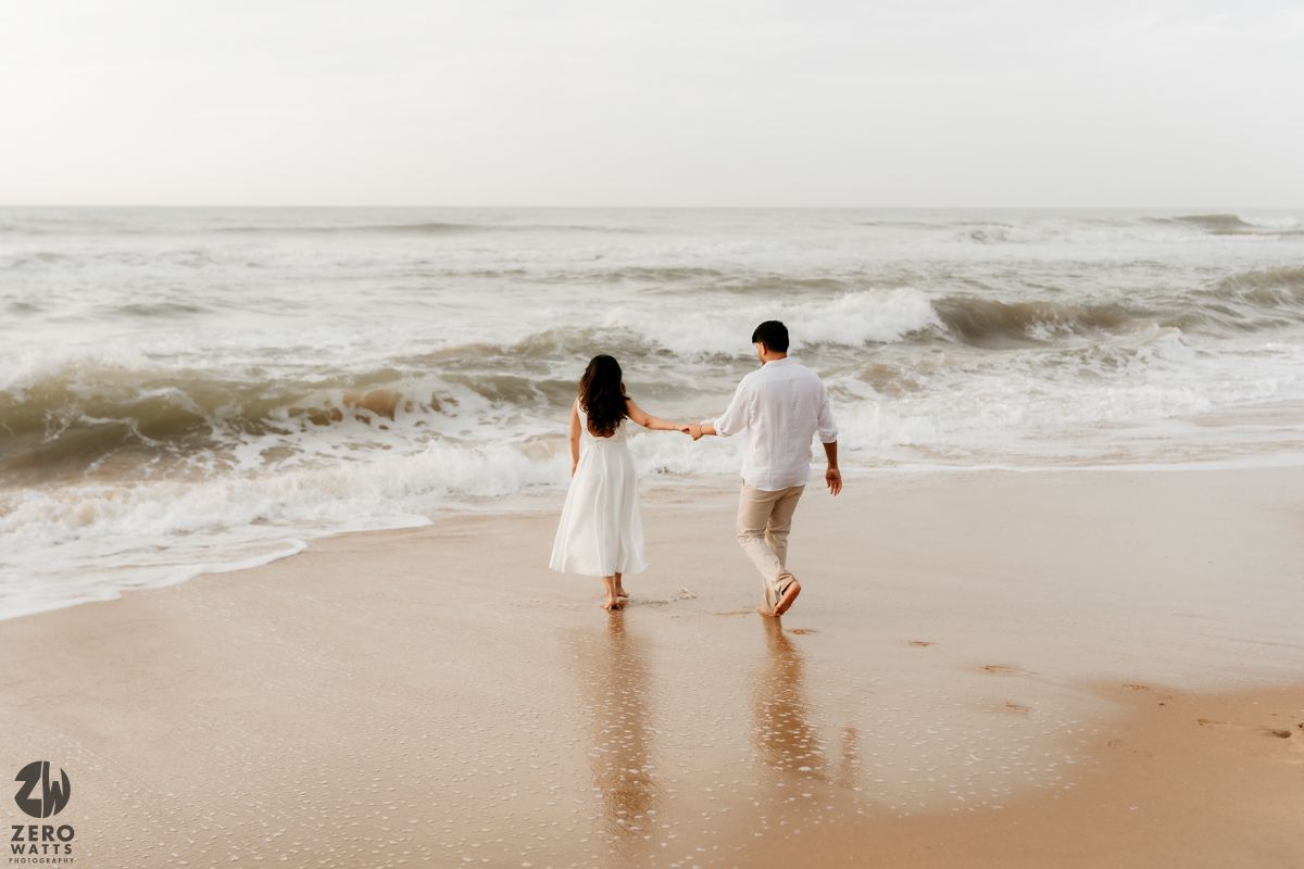Couple walking along beach shoreline in white outfits with waves crashing nearby.