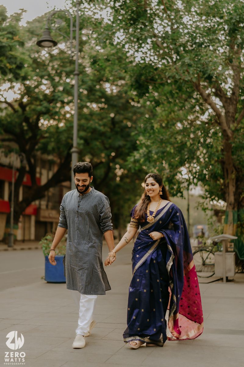 Couple walking hand in hand on a tree-lined path, with a woman in blue saree and man in grey shirt and white pants.