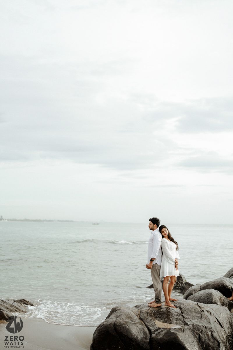 A couple stands on coastal rocks overlooking the ocean on an overcast day.