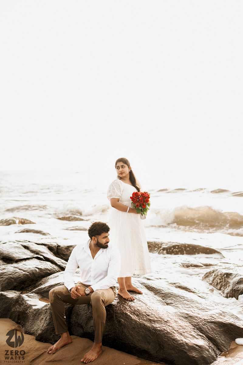 Couple sits on coastal rocks at sunset with waves crashing behind them.