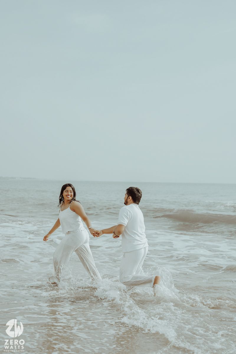 Carefree couple splashing and playing in ocean waves wearing all white outfits.