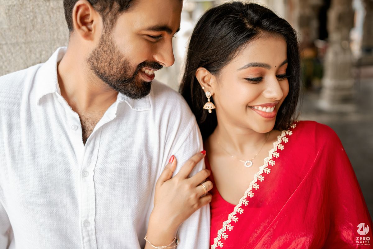 Intimate portrait of couple in white kurta and red saree smiling under temple colonnade.