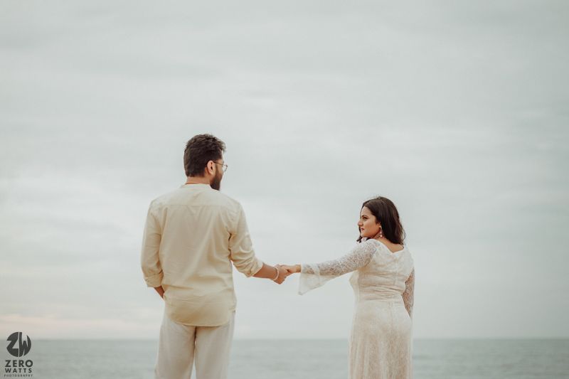 A couple in light-colored outfits holds hands while walking along a misty beach.