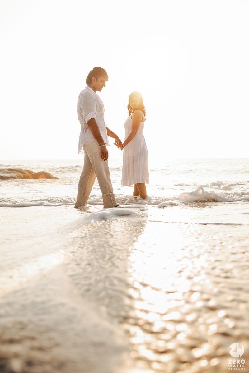 A couple standing in shallow ocean waves at sunset wearing coordinated white and cream outfits.