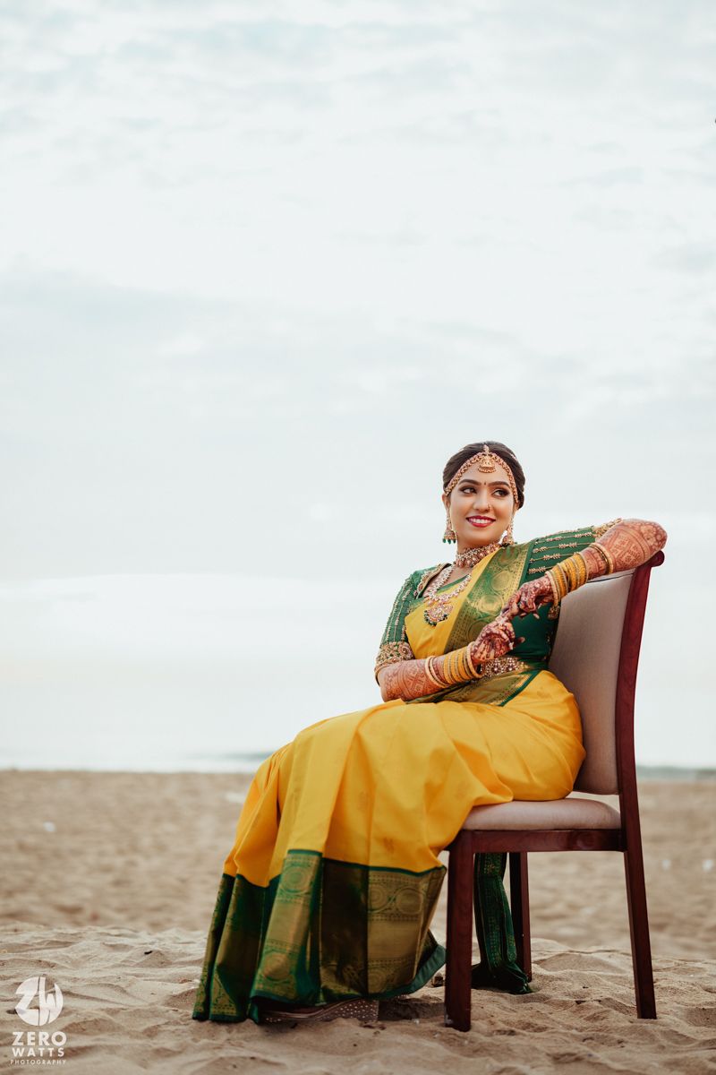 A person in traditional South Indian wedding attire sits gracefully on a wooden chair at the beach.