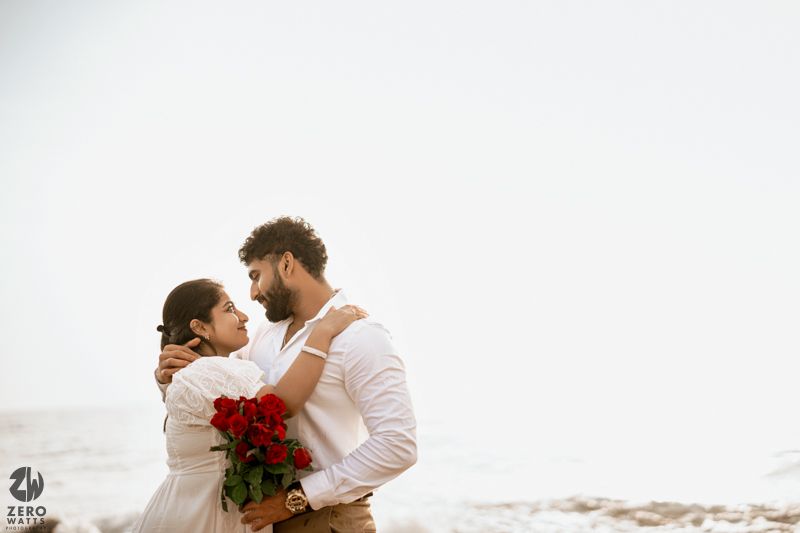 A couple in white clothing poses together on coastal rocks with vibrant red roses.