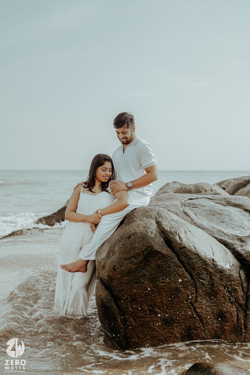 A couple poses together on coastal rocks with the ocean and soft sky in the background.