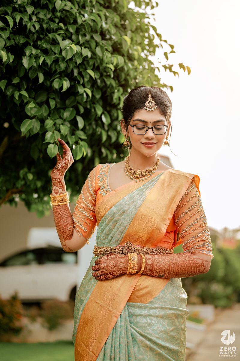 Beautiful Indian bride in mint and orange silk saree with traditional wedding jewelry and henna decorated hands poses elegantly.