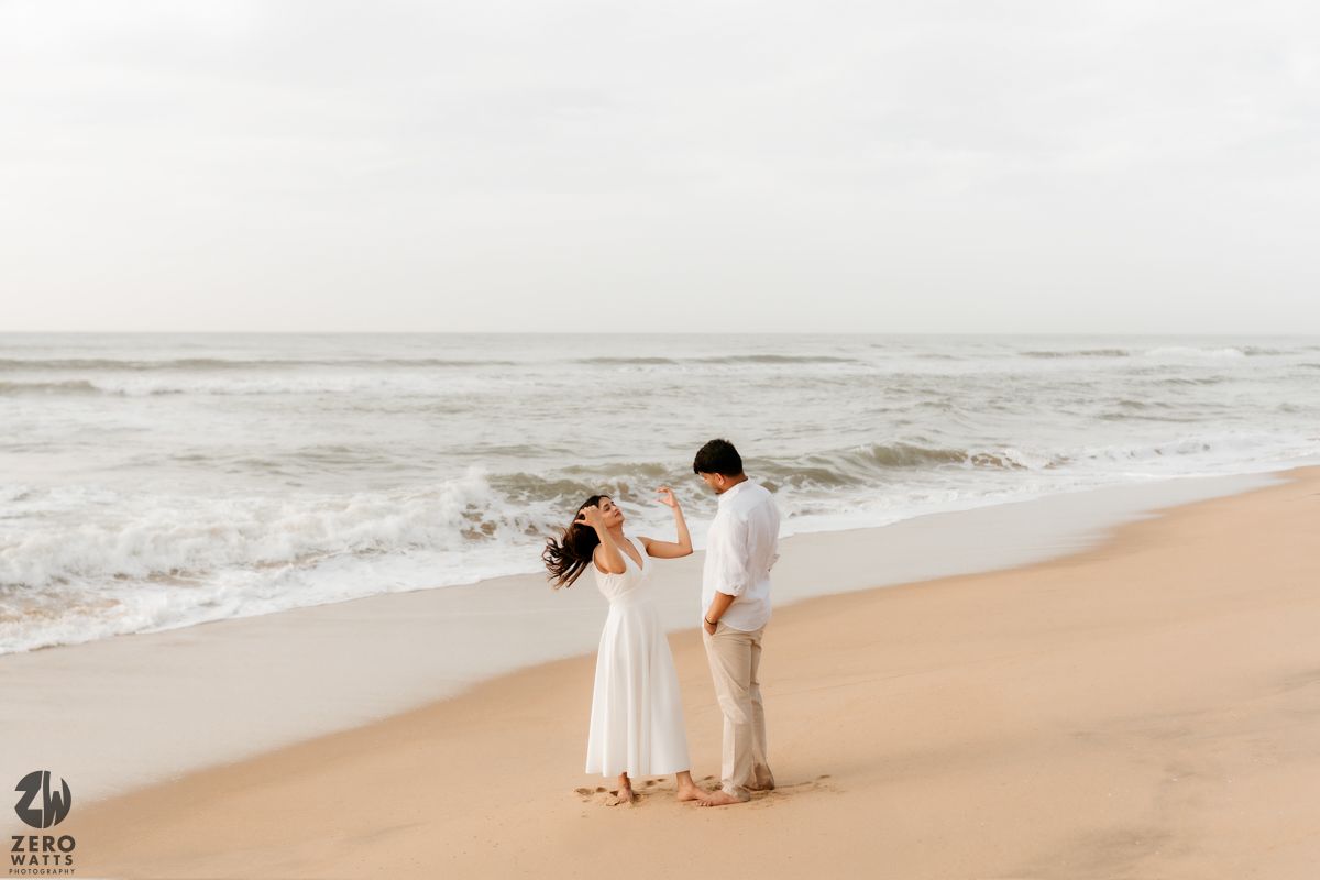 Couple in white outfits walks along misty beach shoreline as waves crash on the sand.