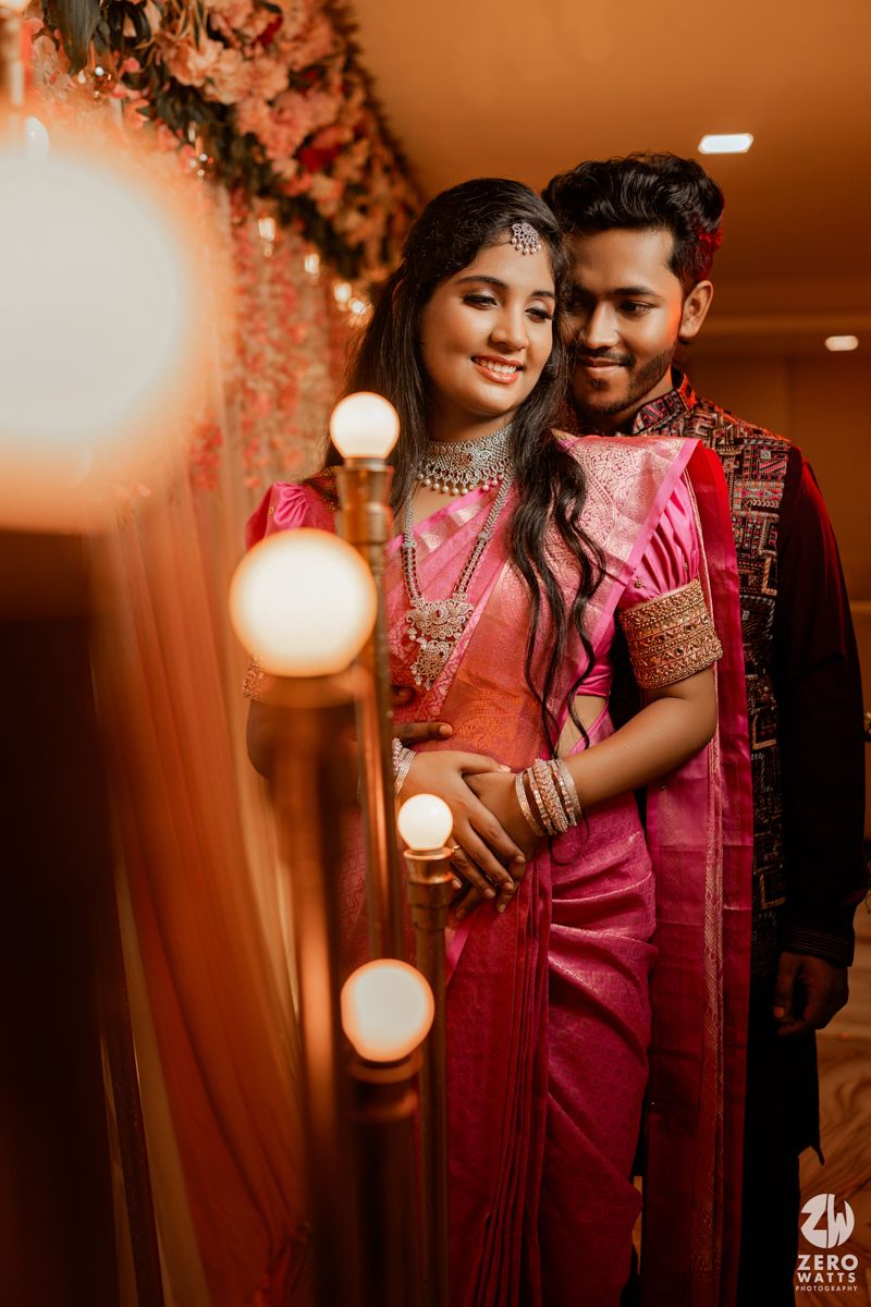 A romantic couple in traditional Indian attire pose together with glowing lights and floral decorations in warm ambient lighting.