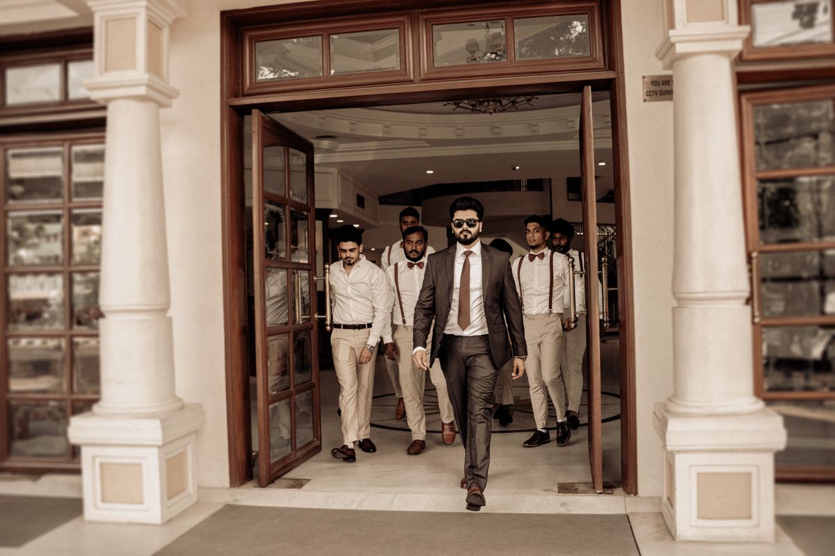 Groomsmen in beige suits and a groom in dark suit walk out through elegant white pillared doorway entrance.