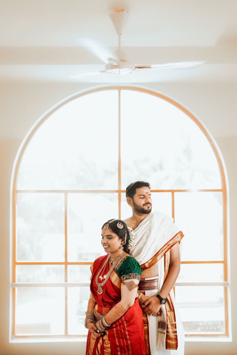 A couple wearing traditional Indian wedding attire stands in front of an arched window with bright natural light.