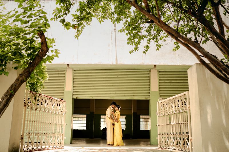 A couple stands in a beautiful entryway with ornate white gates and a green awning framed by leafy tree branches.