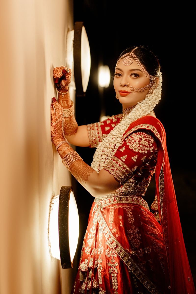 Traditional Indian bride in a red embroidered lehenga and white floral hair accessories poses elegantly near wall lights.