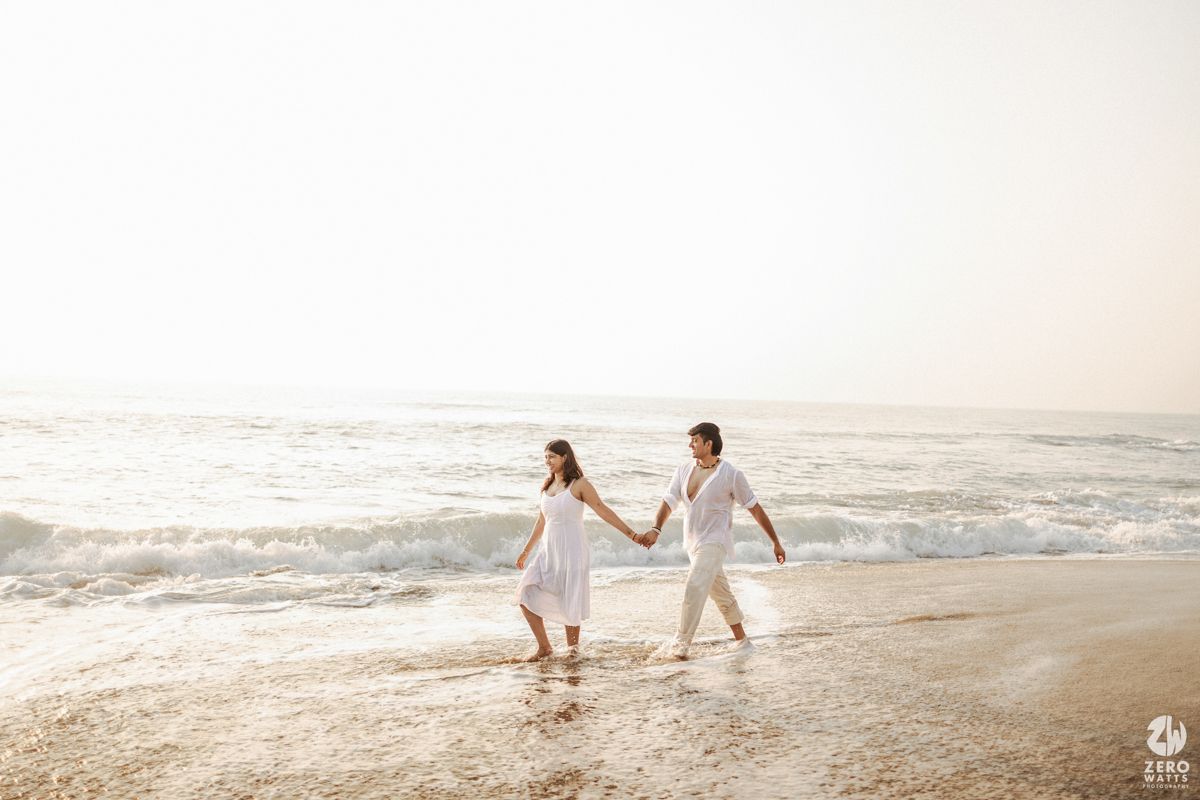 Couple in white clothing runs playfully along beach shoreline during sunset.
