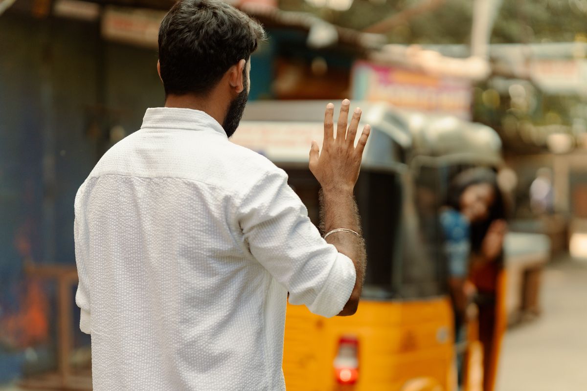 Someone in a white shirt waves goodbye on a blurry city street with an auto rickshaw visible in the background.