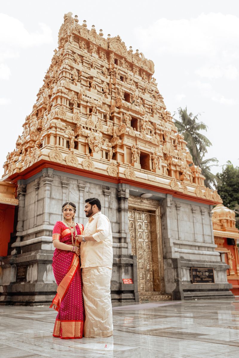 A couple in traditional Indian attire poses in front of an ornate South Indian temple gopuram with intricate carvings.