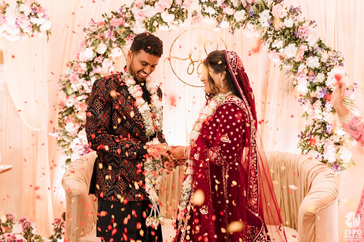 Romantic wedding ceremony under a floral arch with rose petals falling in soft pink lighting.
