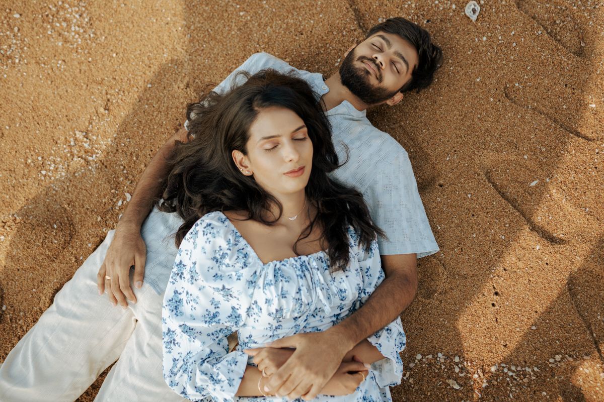A couple lies on sandy beach together wearing matching light colored outfits.