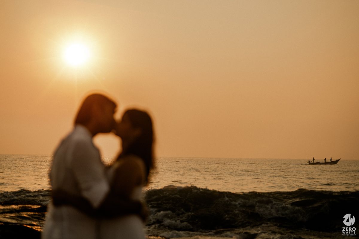 Silhouetted couple sharing an intimate moment during sunset at the beach with a small boat on the horizon.