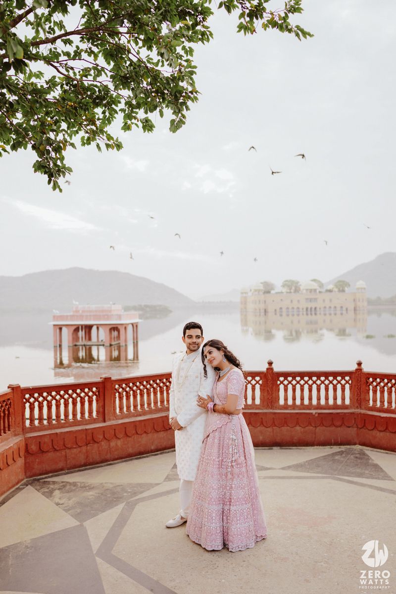 Couple poses elegantly on a red terrace overlooking a misty lake with a palace in the background.