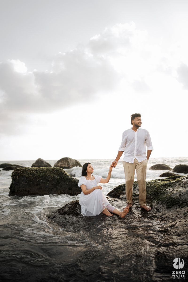 A couple shares a romantic moment on rocky coastal cliffs during sunset with cloudy skies in the background.
