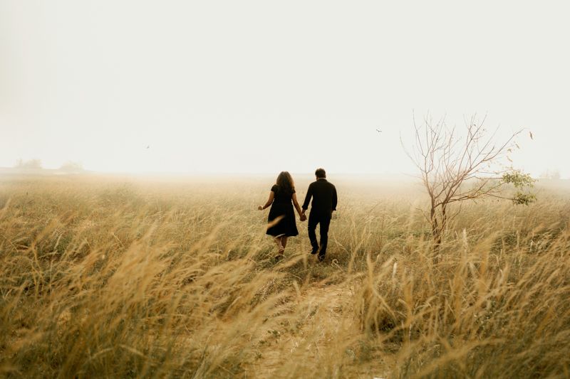 Two silhouetted figures walk together through tall golden grass in a misty field during sunset.