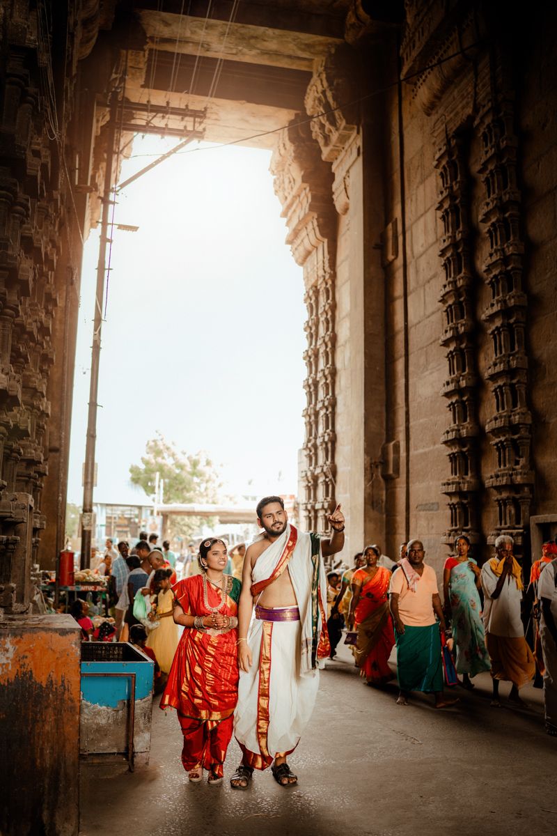 Traditional Indian couple stands in ornate temple corridor with carved pillars as visitors pass by in warm sunlight.