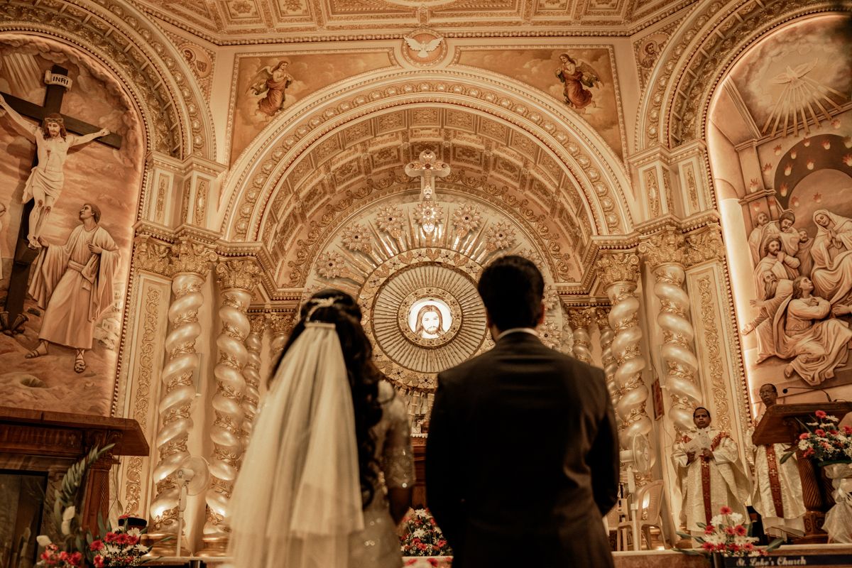 A couple stands in a grand cathedral with ornate baroque architecture, illuminated by warm lighting during a wedding ceremony.