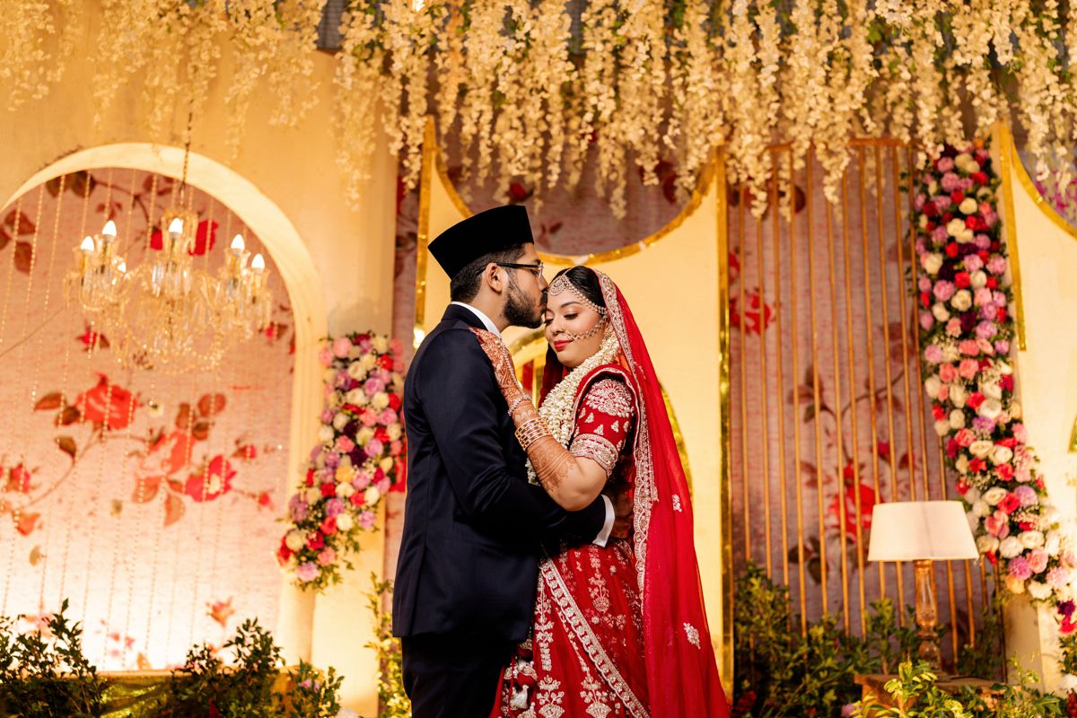 A newlywed couple in traditional South Asian wedding attire embraces under a floral archway with hanging decor.
